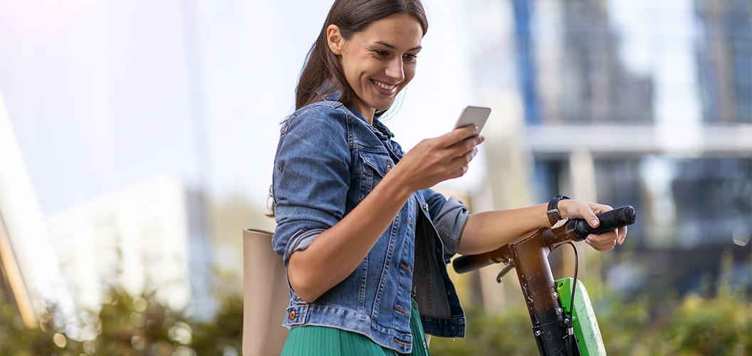 Persona apoyada en un patinete eléctrico mientras utiliza un teléfono móvil en un entorno urbano