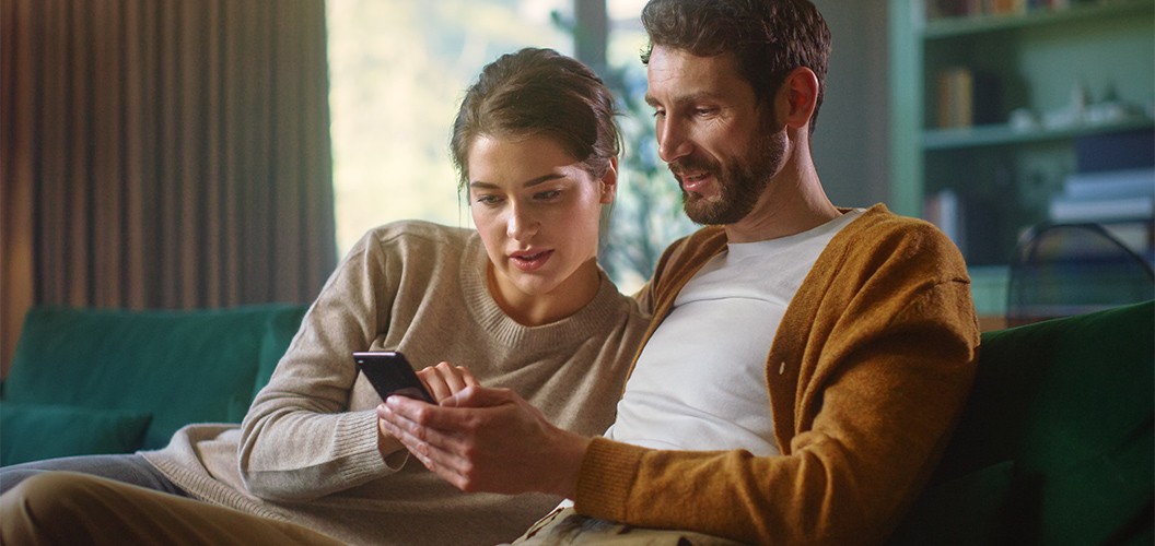 Dos personas sentadas en un sofá revisando información en un teléfono móvil dentro de un salón iluminado y con estanterías al fondo